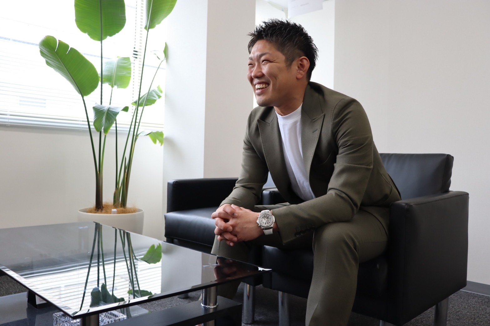 緑色のスーツを着た男性が、モダンなオフィスの黒いソファに笑顔で座っており、近くには大きな鉢植えとガラスのコーヒーテーブルが置かれている。.