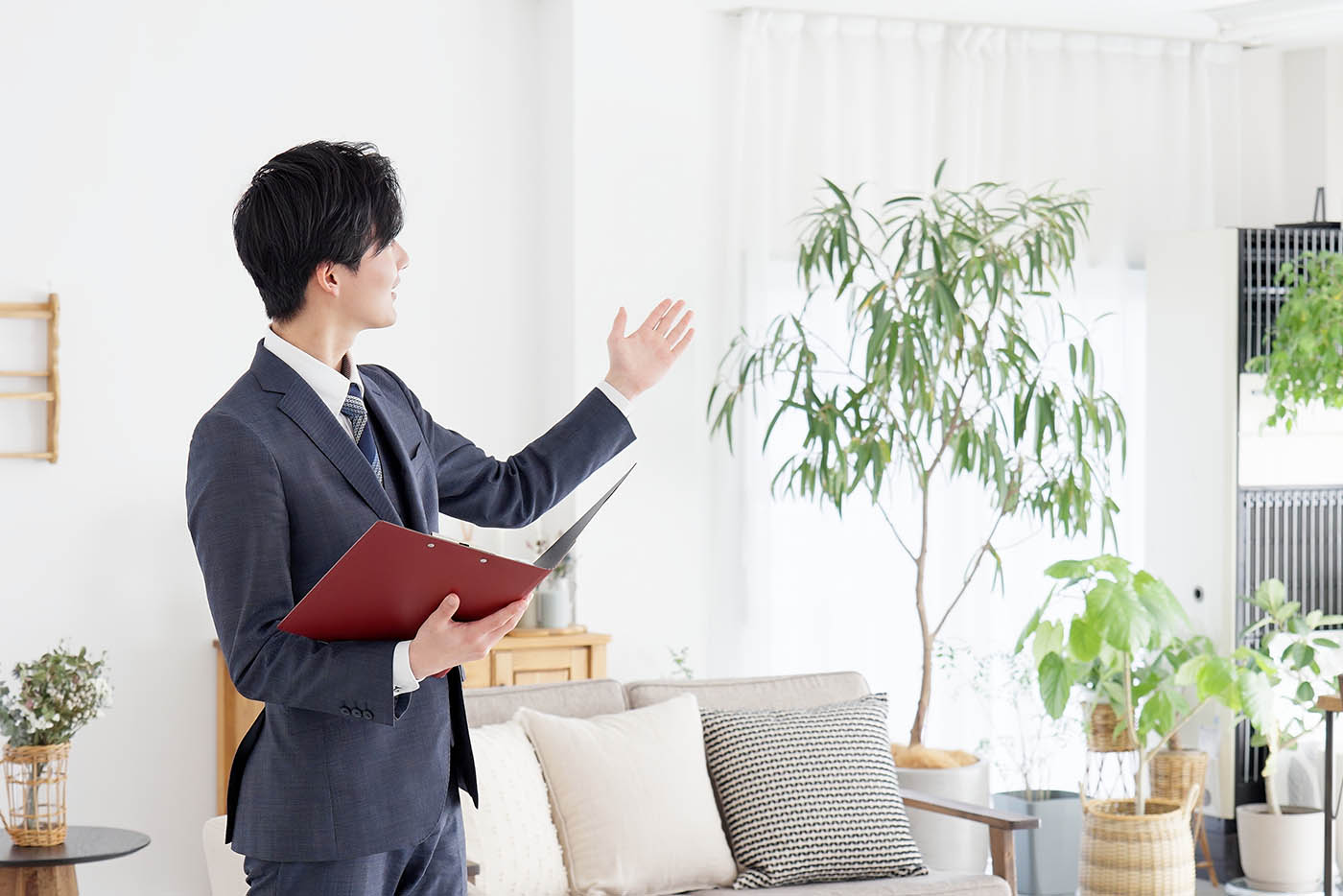 赤いフォルダーを持ったスーツを着た男性が、観葉植物と中間色の家具が置かれた明るくモダンなリビングルームを指しています。.