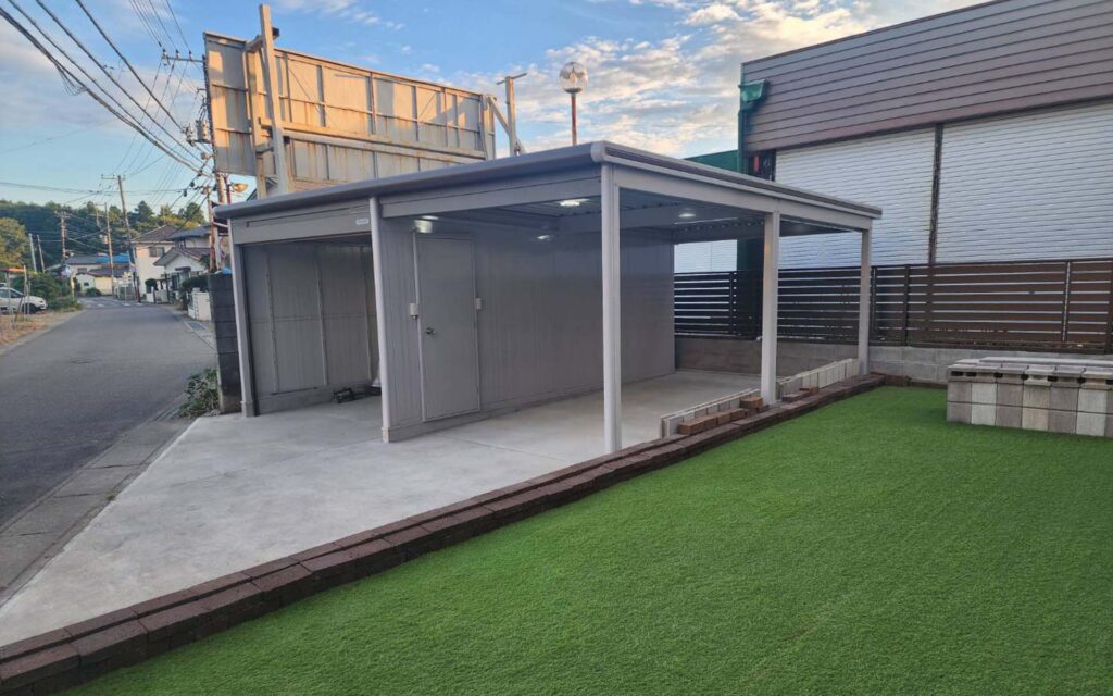 曇り空の下、住宅街の屋外エリアにある人工芝の横のコンクリートスラブの上に、モダンな金属製のカーポートが立っています。背景には道路、フェンス、建物が見えます。.