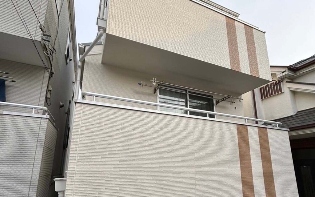 縦縞のベージュが特徴的な2階建ての白いモダンな建物。金属製の手すりが付いたバルコニーと大きな正面窓が特徴です。隣接する建物の間に位置しています。.