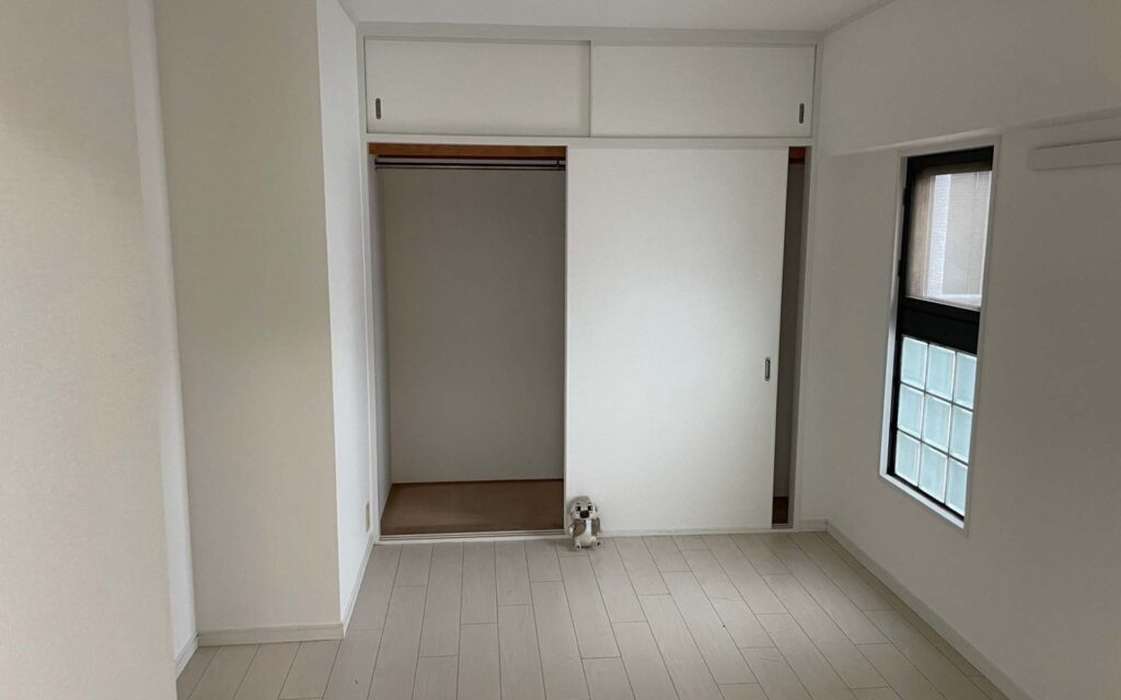 明るい色の空っぽの部屋。床はフローリングで、引き戸付きのオープンクローゼットがあり、クローゼットの前の床には小さなぬいぐるみかおもちゃが置かれている。右側の壁には窓がある。.
